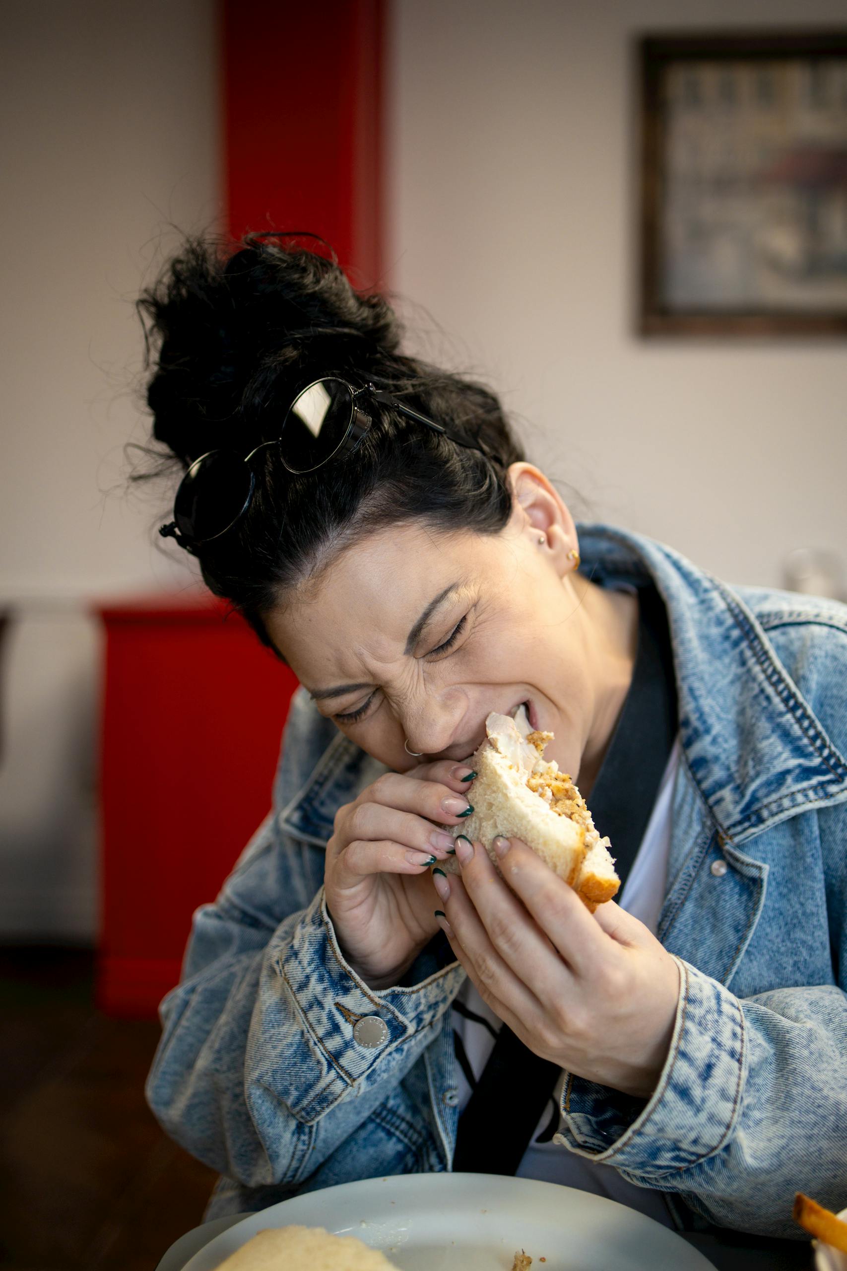A woman in denim jacket joyfully eating a sandwich indoors, symbolizing comfort food.