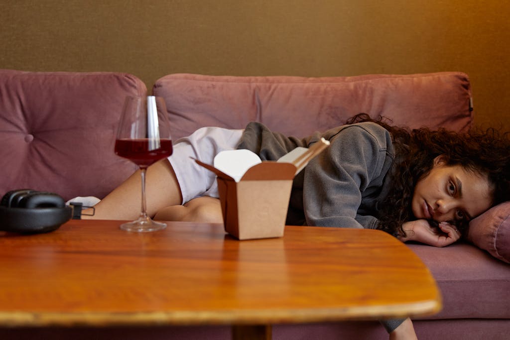 A woman lying on a sofa with a wine glass and takeaway box, reflecting indoors.