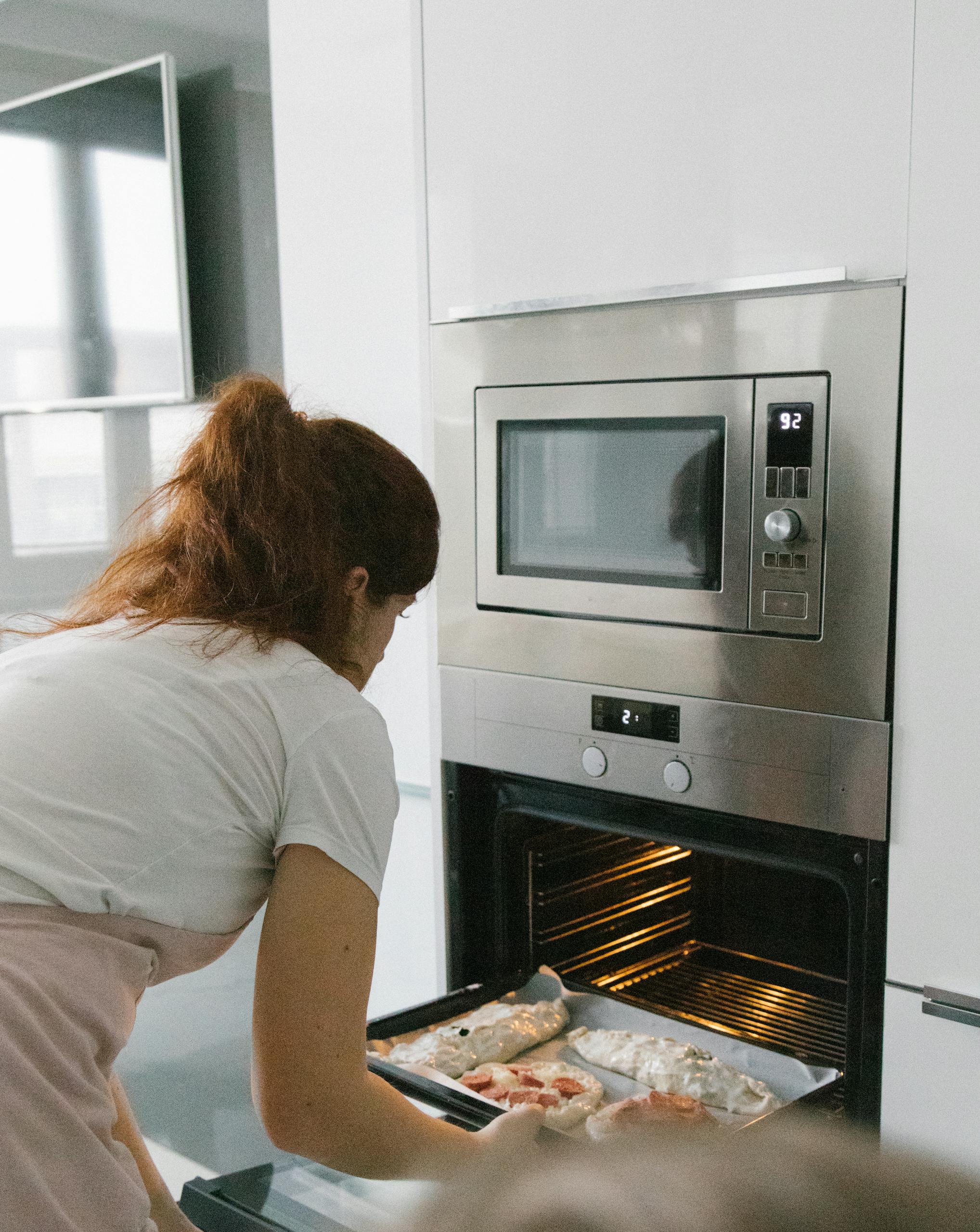 A woman placing a baking tray into an oven while cooking a homemade dish in a contemporary kitchen setting.
