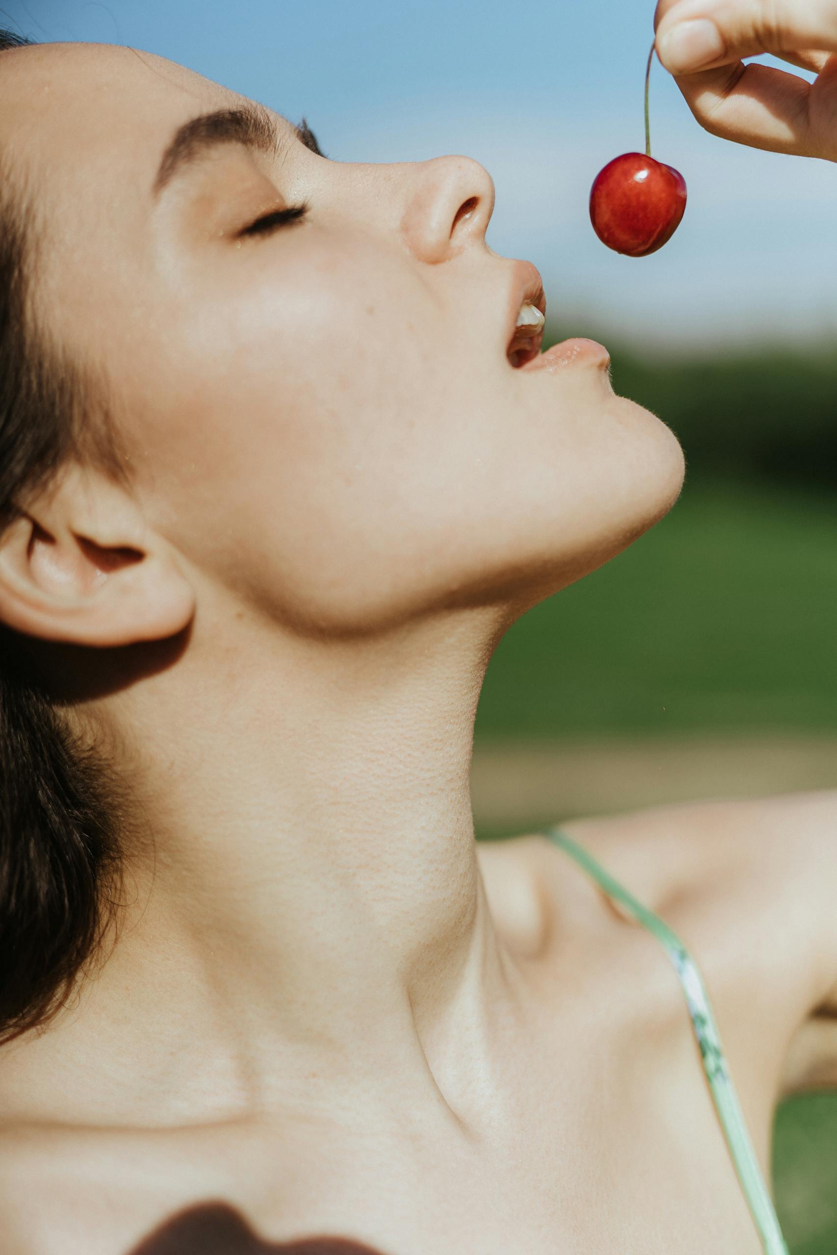 A woman poses under a clear sky, savoring a ripe cherry. Perfect for summer lifestyle and food themes.