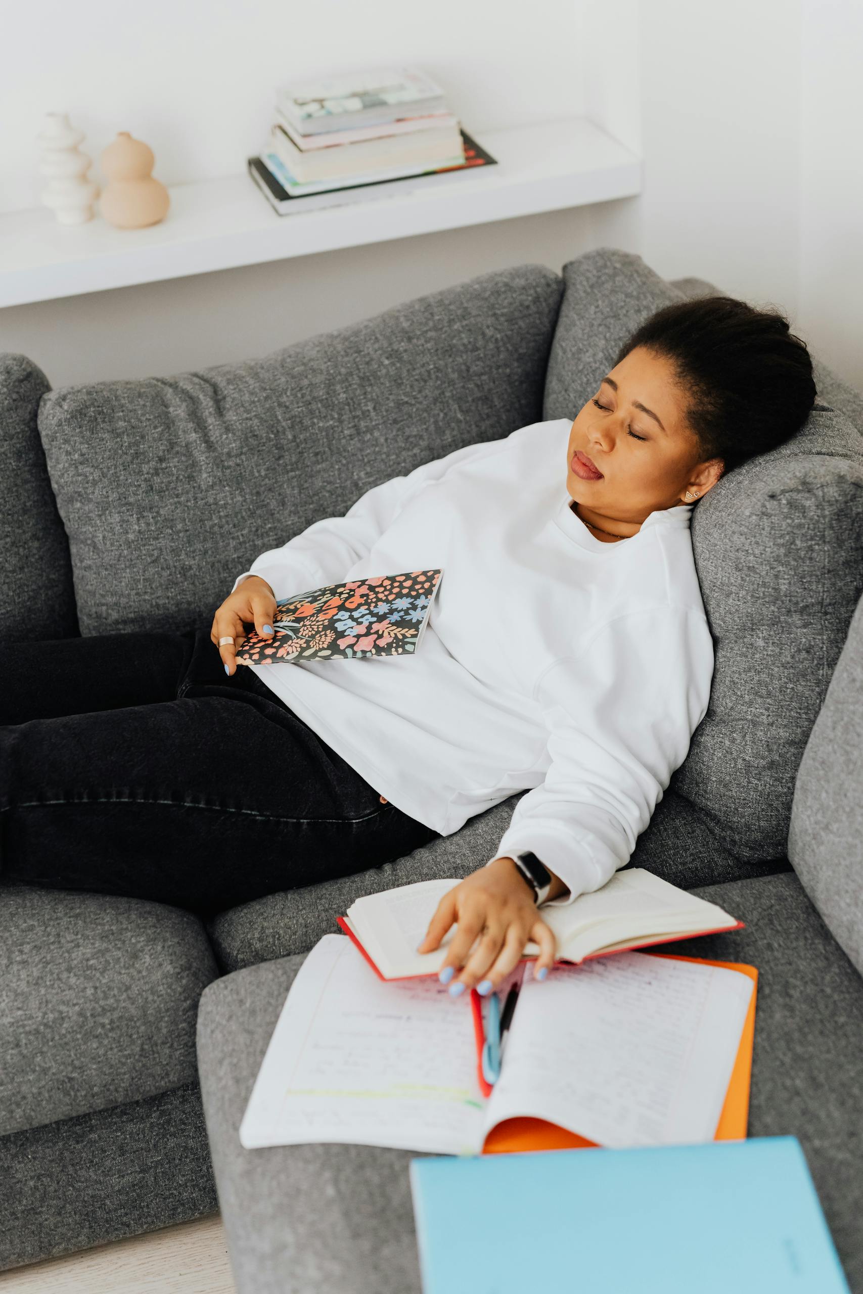 A woman resting on a couch with study materials scattered around, showing academic fatigue.