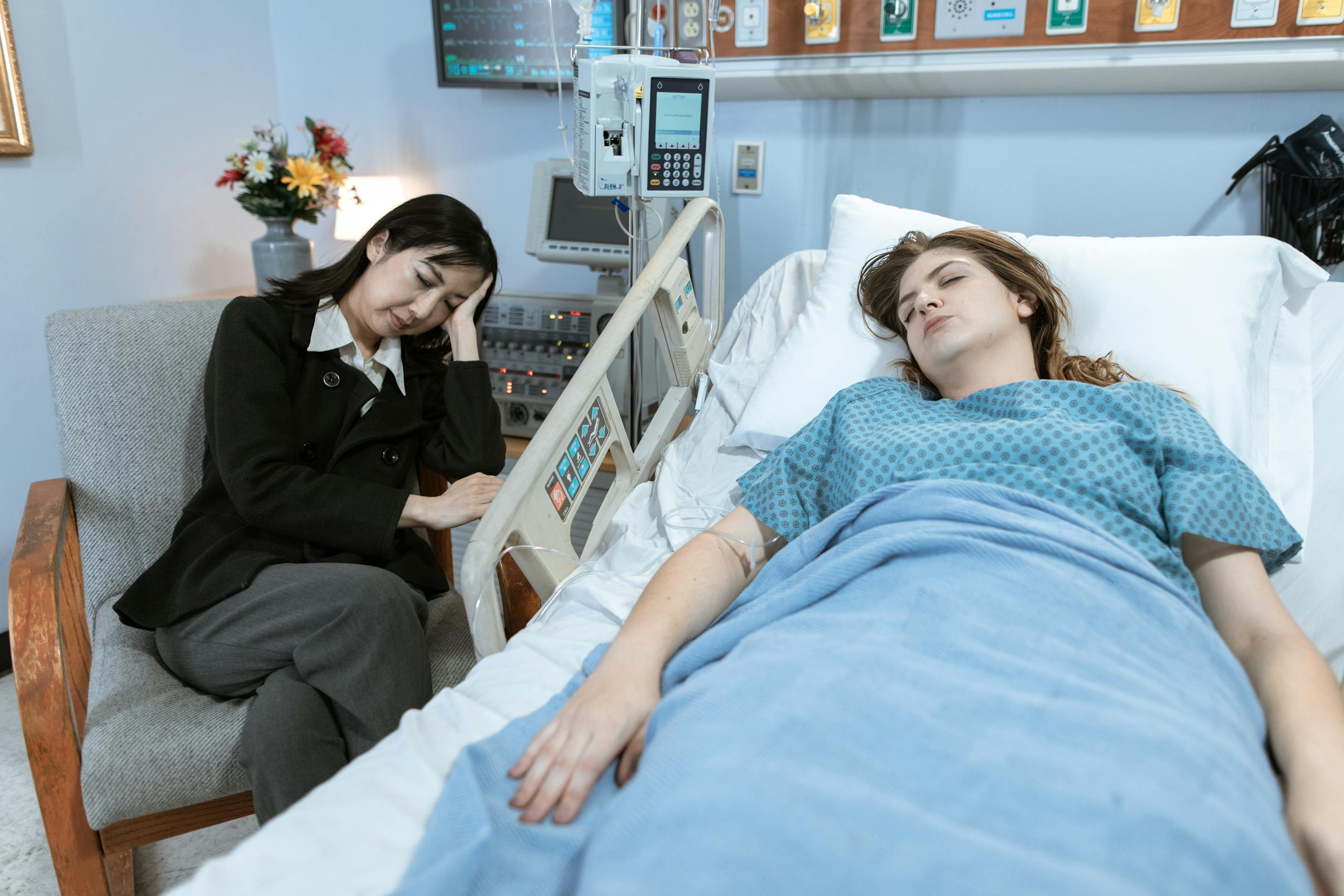 A woman sits by a hospital bed, looking worried, as a patient rests peacefully.