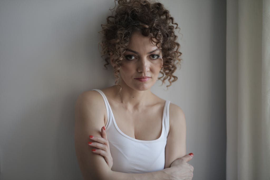 A woman with curly hair in a white tank top stands with arms crossed, exuding a fierce expression.