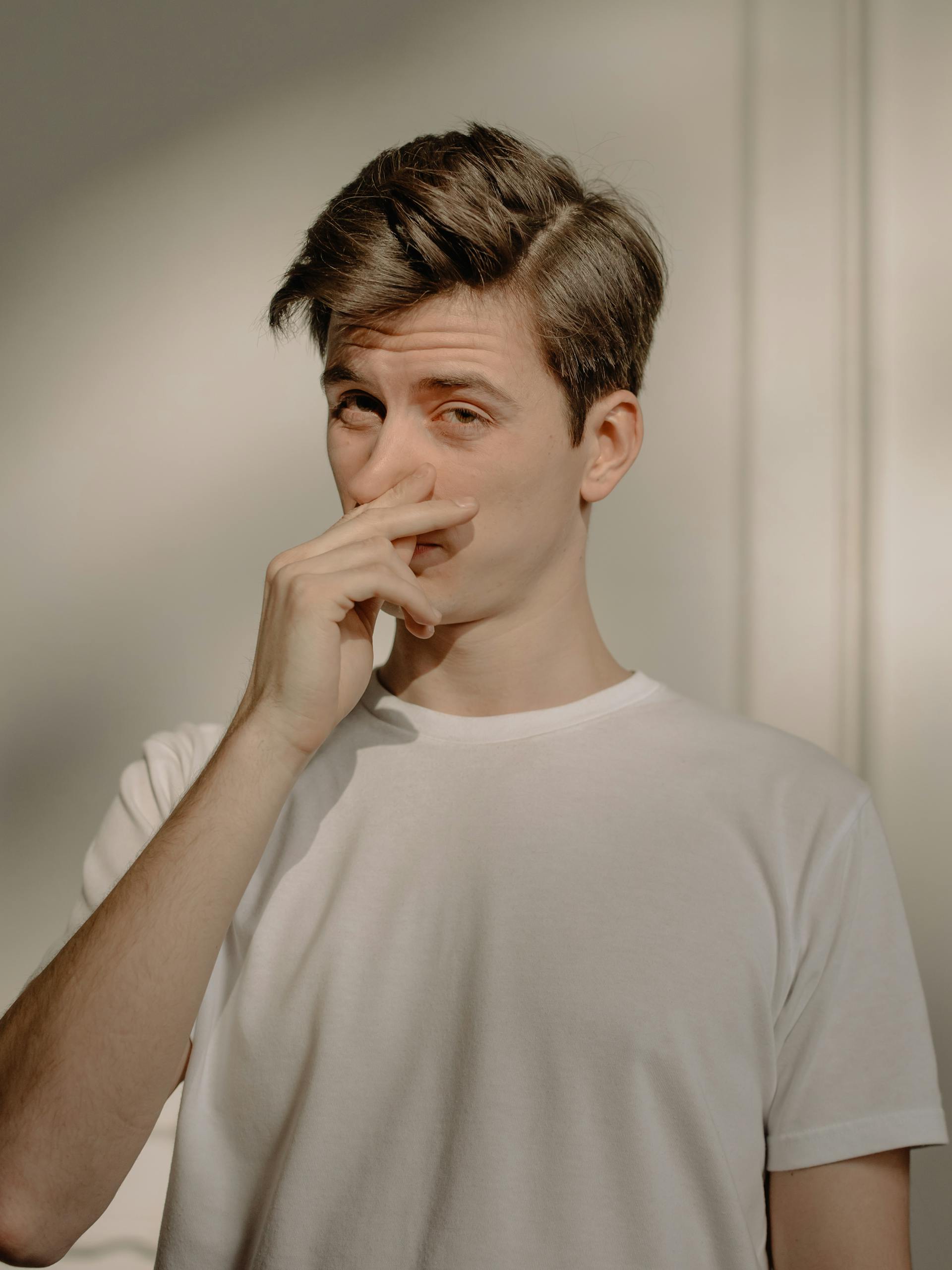 A young man stands indoors in a white T-shirt, covering his nose with a thoughtful expression.