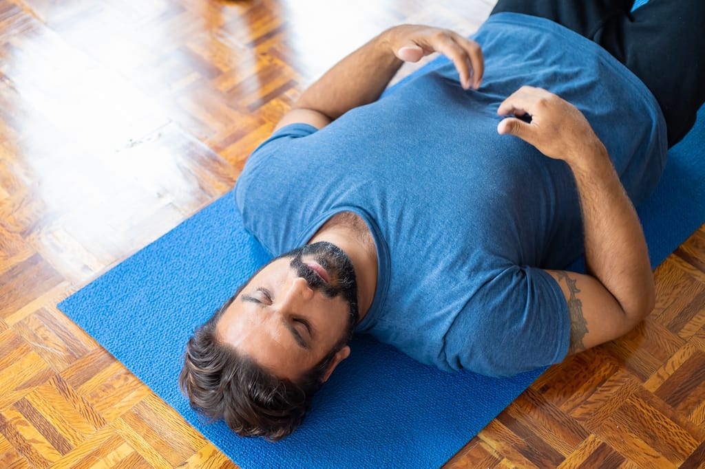 Adult man lying on a blue yoga mat indoors with eyes closed, focusing on relaxation and mindfulness.