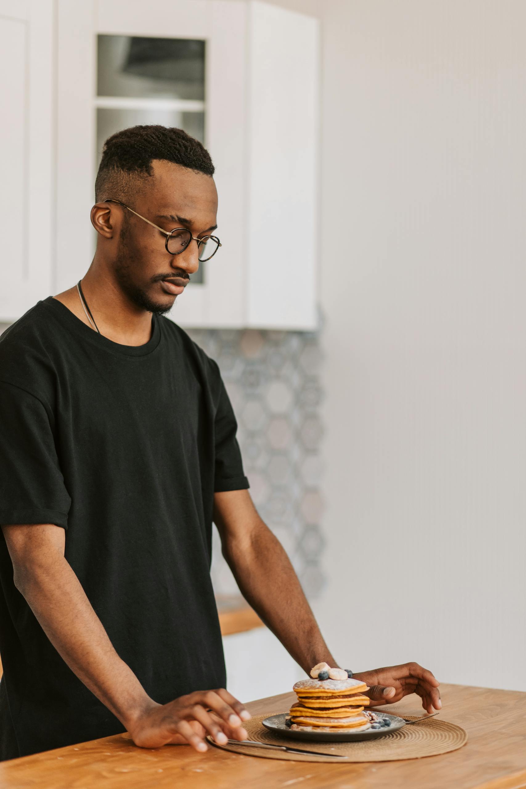 African American man with glasses stands near a fresh stack of pancakes in a modern kitchen.