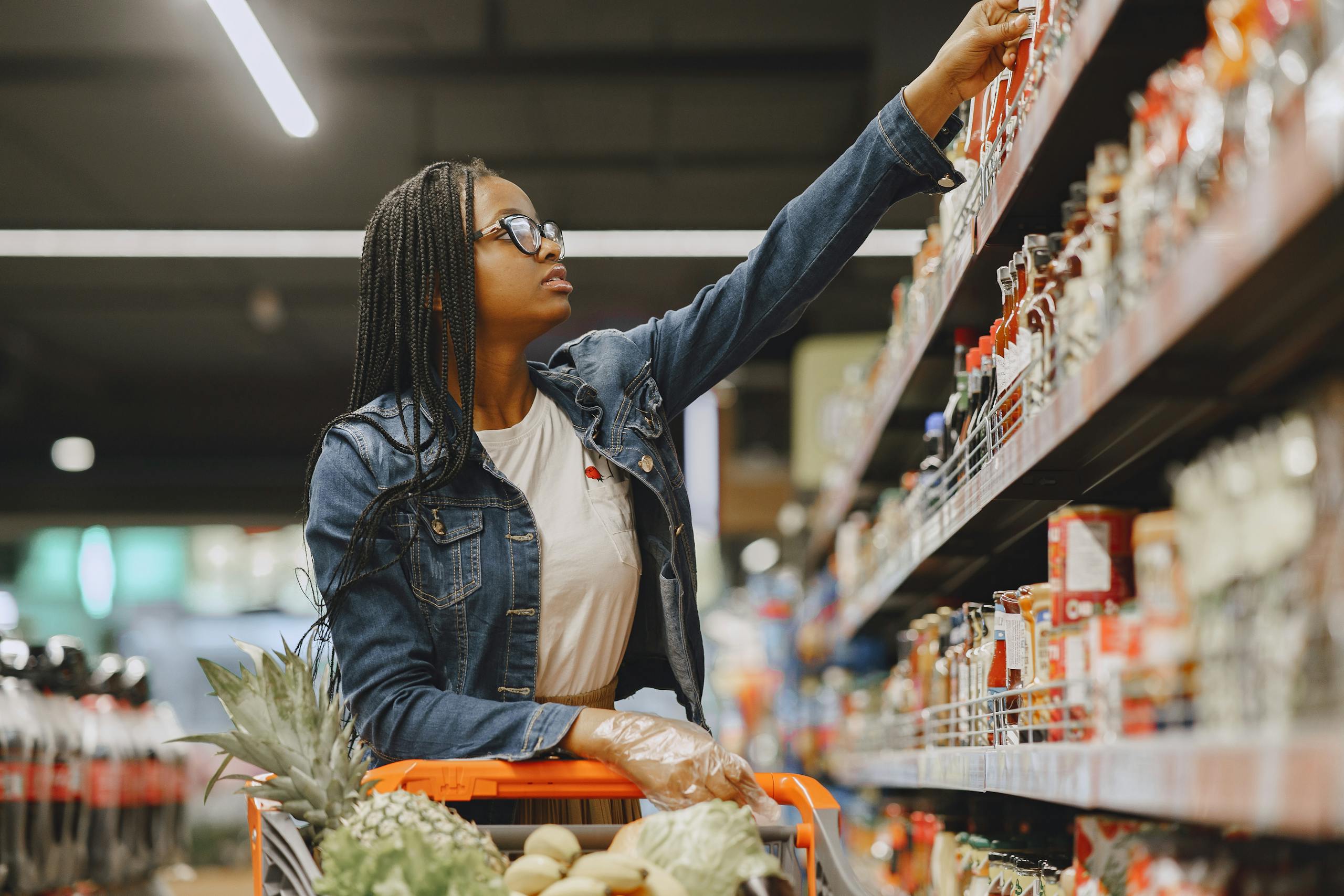 African American woman selecting items in a grocery store aisle with a shopping cart.