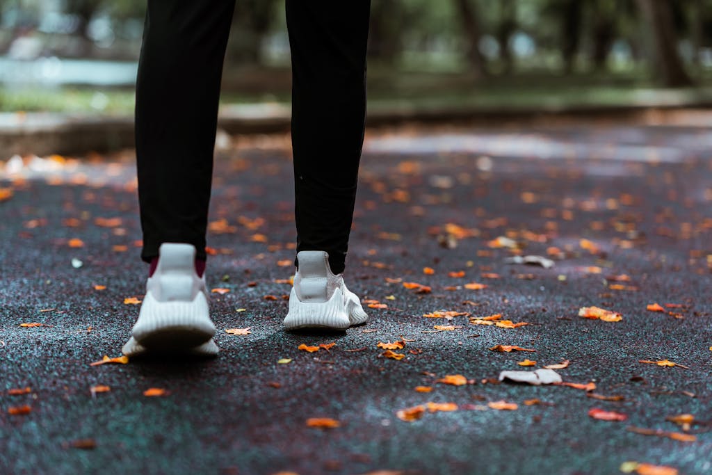 Back view of crop unrecognizable female in leggings and footwear strolling on asphalt road covered with faded leaves in city park in fall