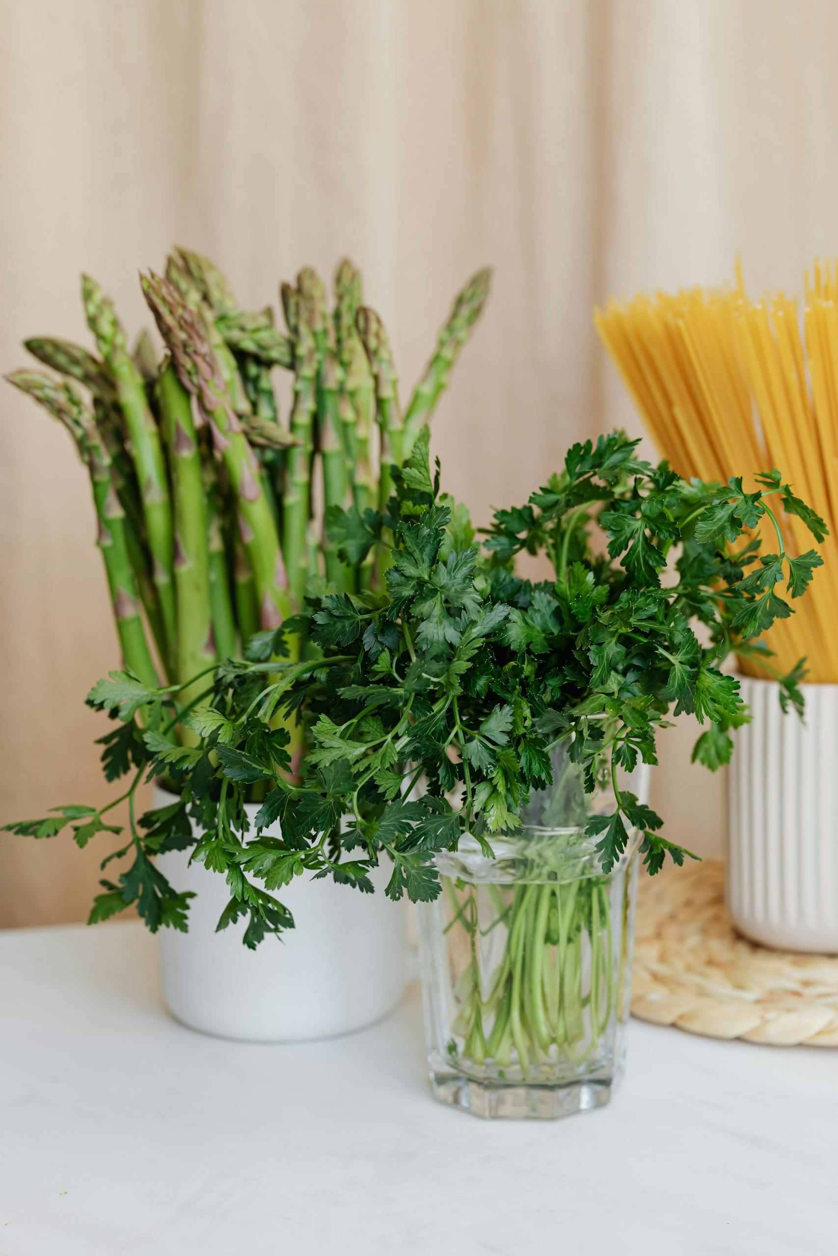 Bouquet of green fresh parsley in glass before cup with green asparagus near yellow uncooked spaghetti in ceramic vase on white table