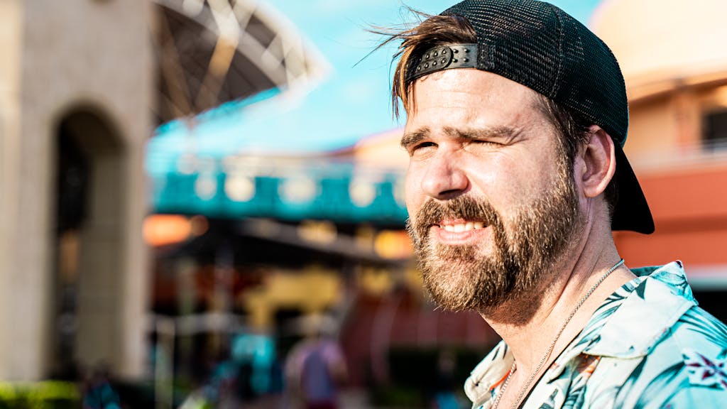 Casual bearded man wearing cap and floral shirt in a sunny urban area.
