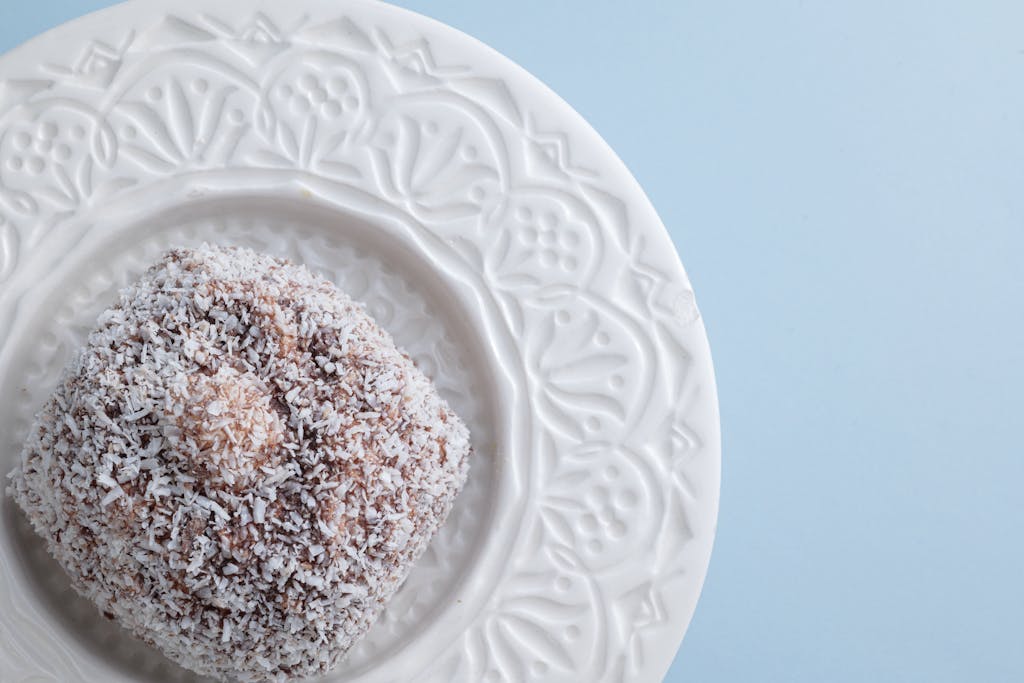 Close-up of a coconut-covered lamington on a patterned white plate on blue background.