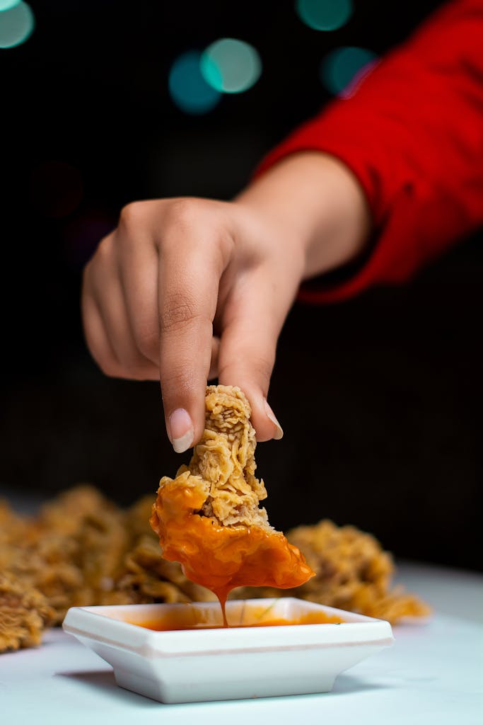 Close-up of a hand dipping a piece of crispy fried chicken into sauce, showcasing texture and deliciousness.