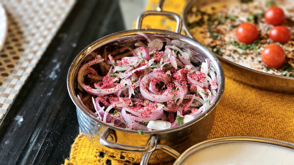 Close-up of a spiced onion salad in a brass bowl, garnished with sumac and herbs, on a vibrant table setting.