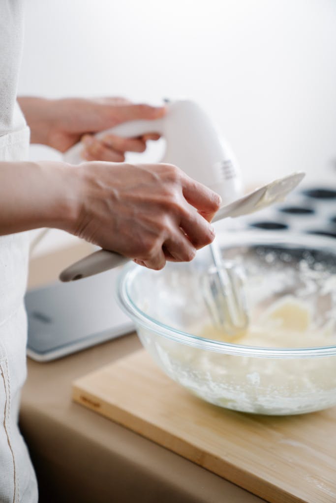 Close-up of hands using an electric mixer to mix dough in a glass bowl on a wooden board.