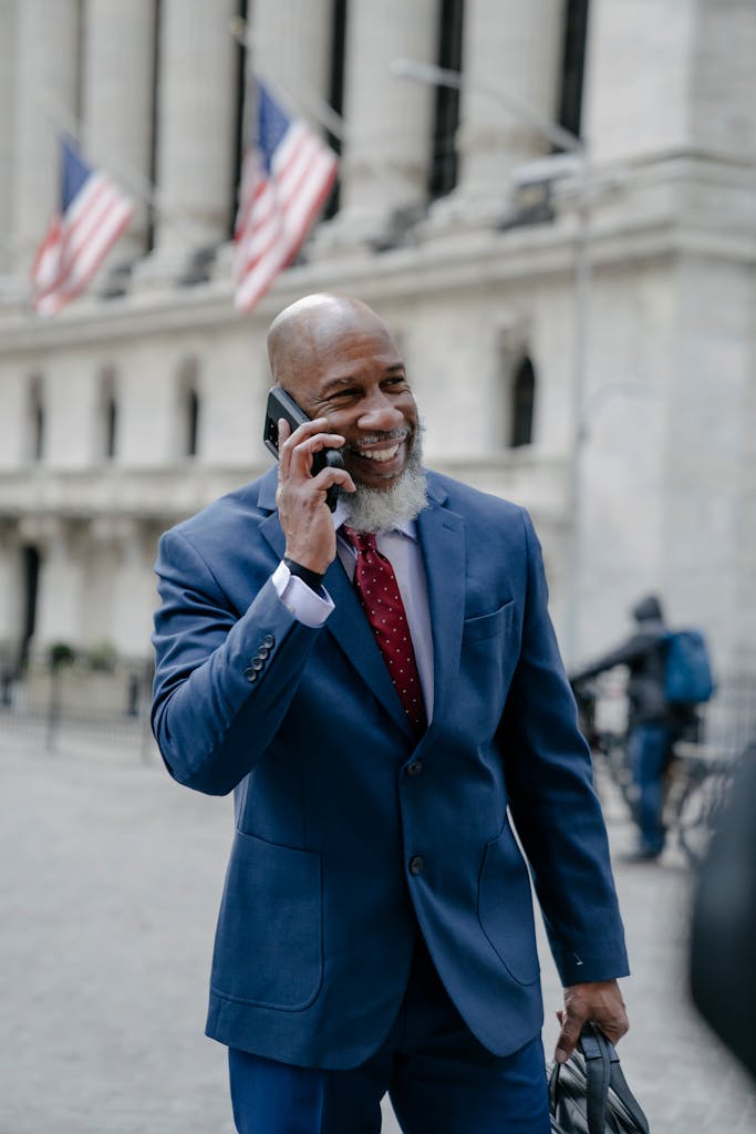 Confident bald man in blue suit making a phone call outside an iconic building with flags.
