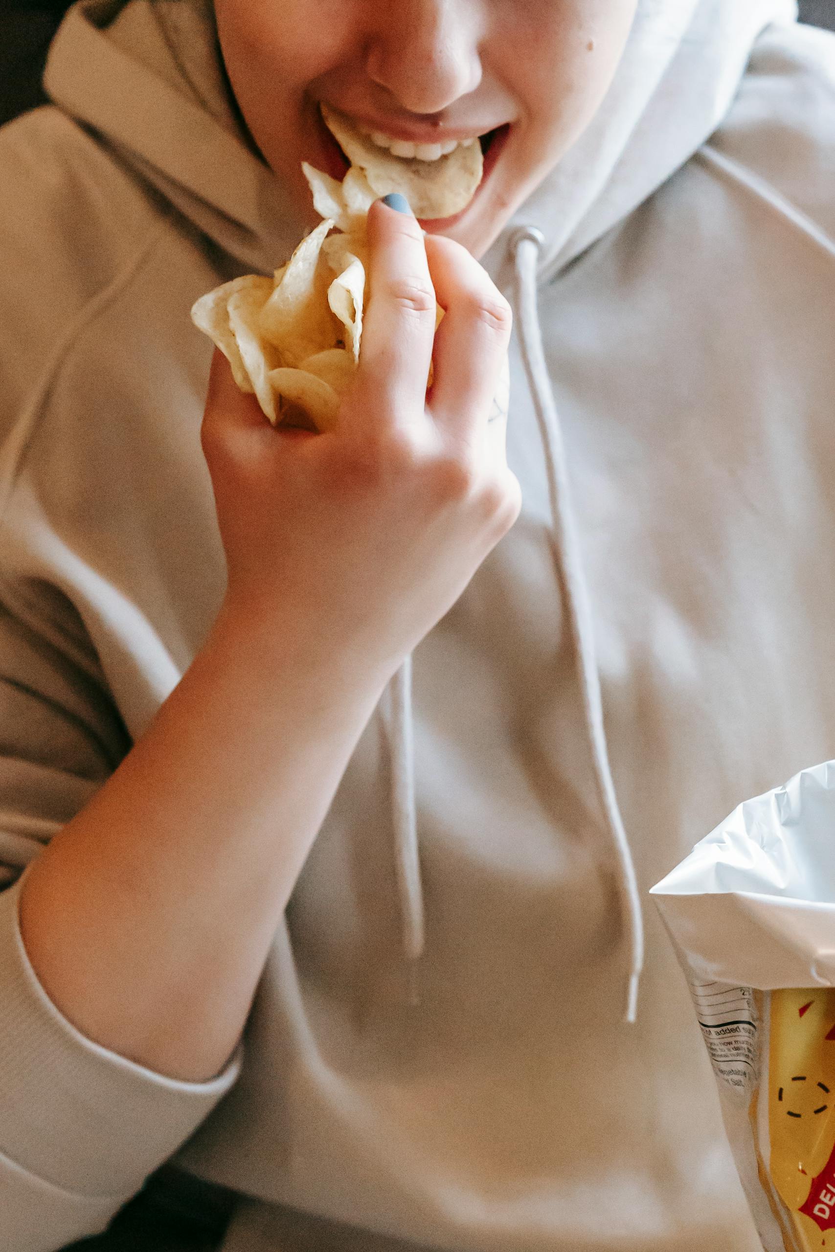Crop female in comfy hoodie eating tasty savory potato chips in light room