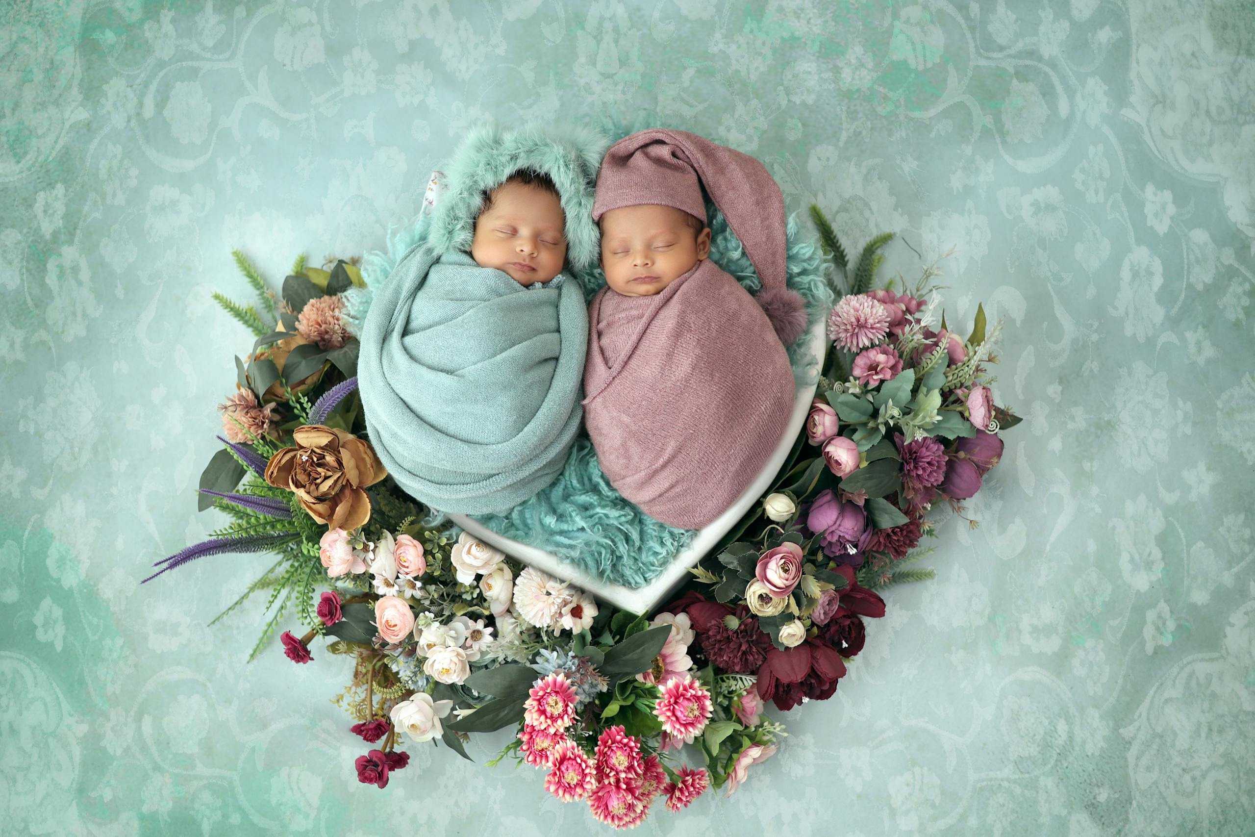 Cute twin babies wrapped in pastel swaddles, nestled in a floral arrangement.