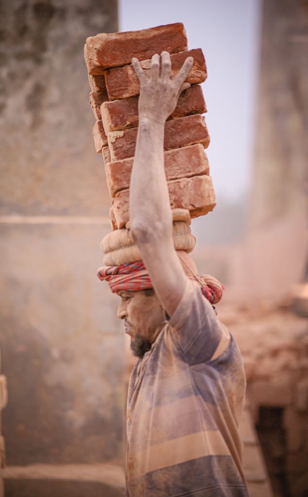 Dedicated worker transports bricks on head at dusty construction site, showcasing strength and resilience.