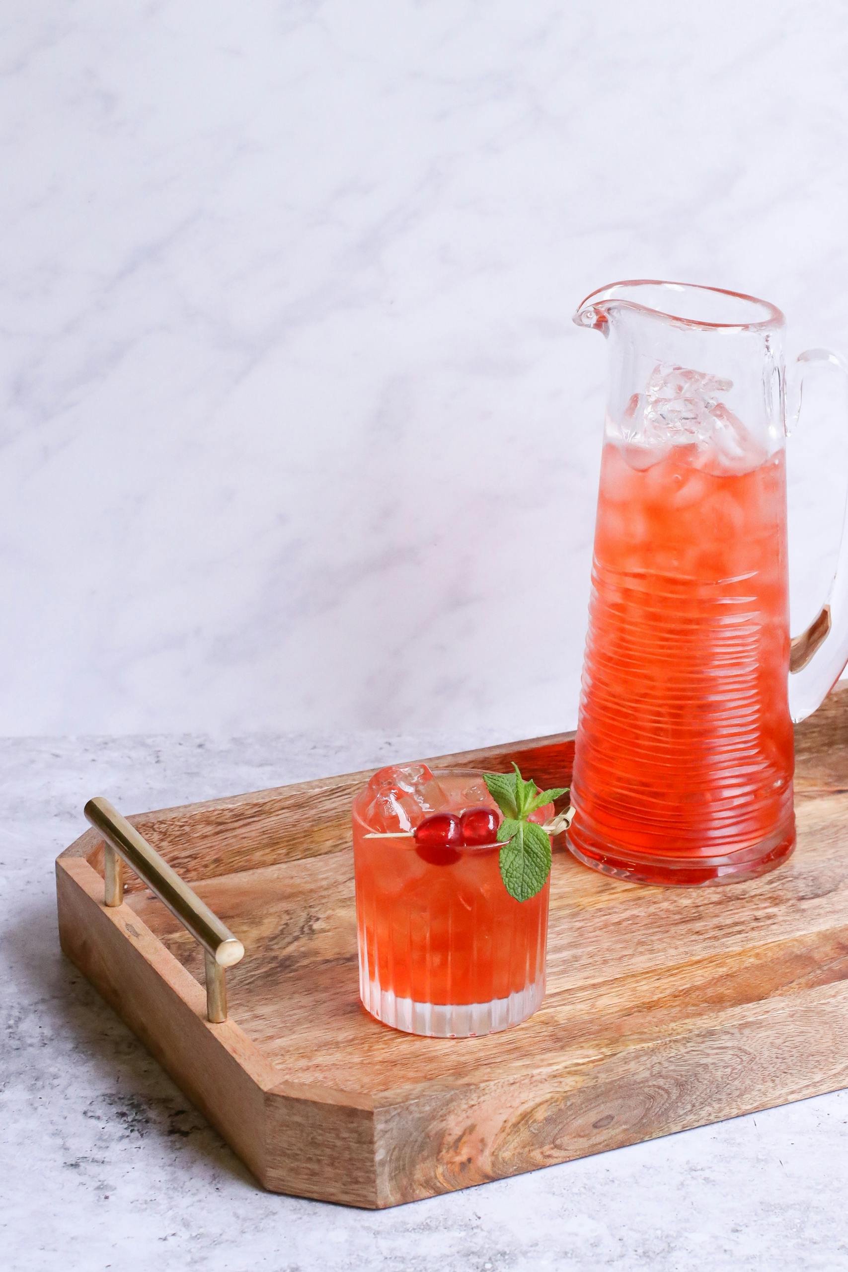 Delicious beverage with ice and fresh cherries near decanter on wooden tray on light background