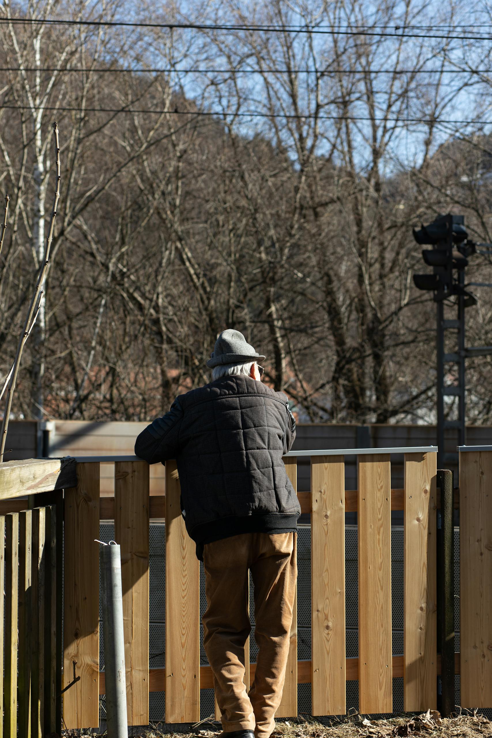 Elderly man in black jacket stands by a wooden fence, contemplating winter landscape.