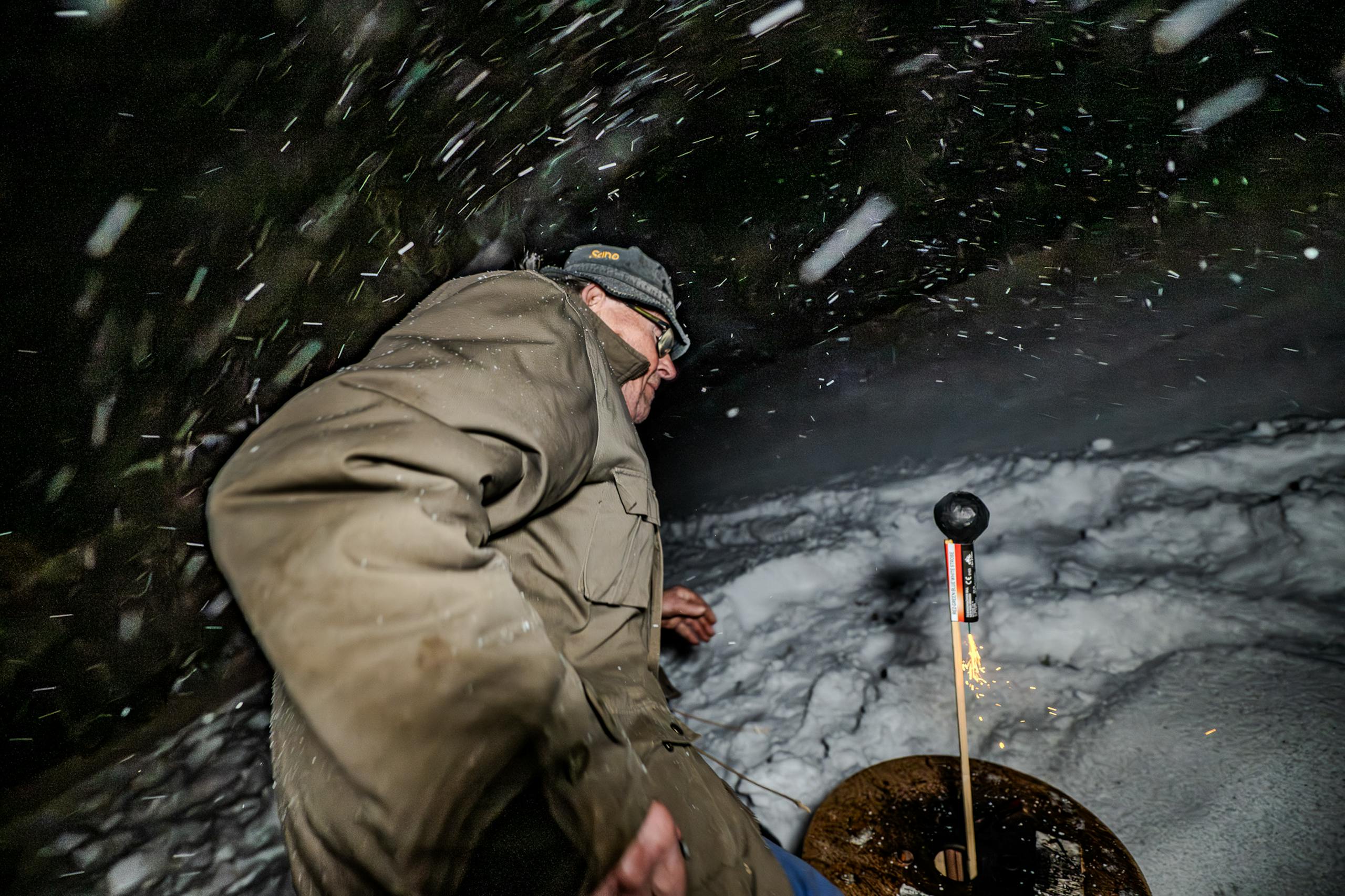 Elderly man lights a firework in snowy outdoor night setting, sparks flying.