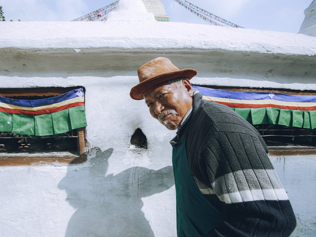 Elderly man wearing a brown hat and sweater standing by a traditional white structure.
