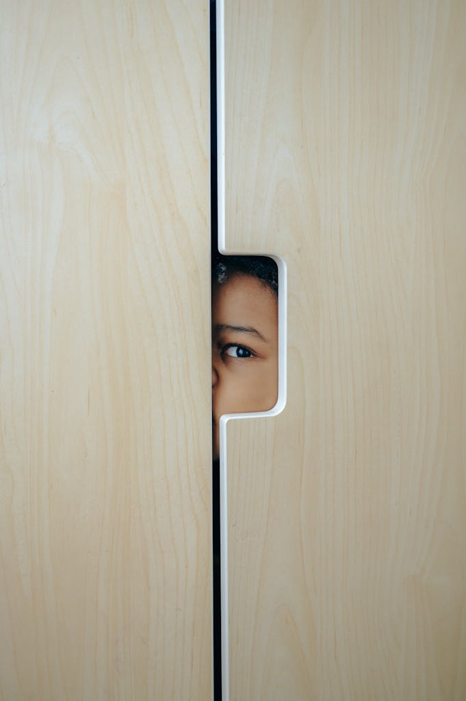 Eye of unrecognizable funny little ethnic child hiding in wooden wardrobe and looking through hole during funny game