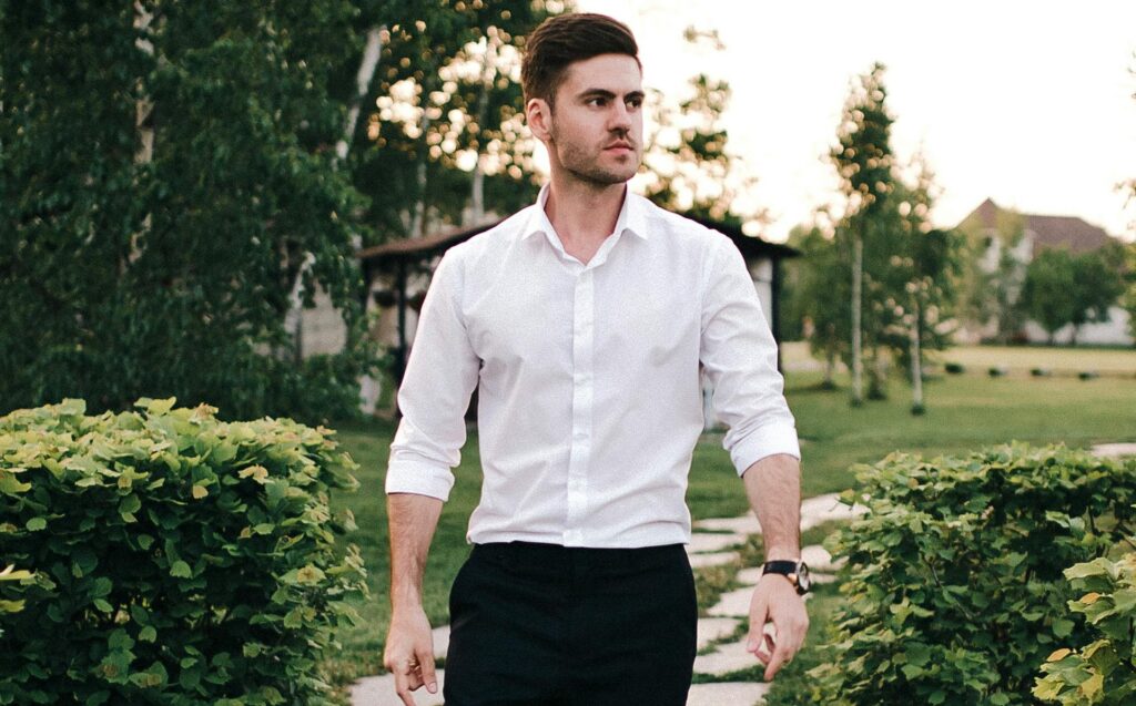 Fashionable man in a white shirt and black pants walking on a wooden path in a green park setting.