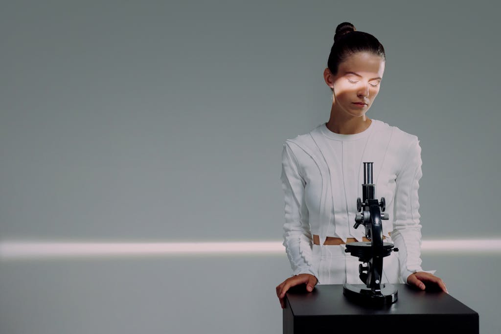 Female scientist in white illuminated by light next to a microscope in a studio.