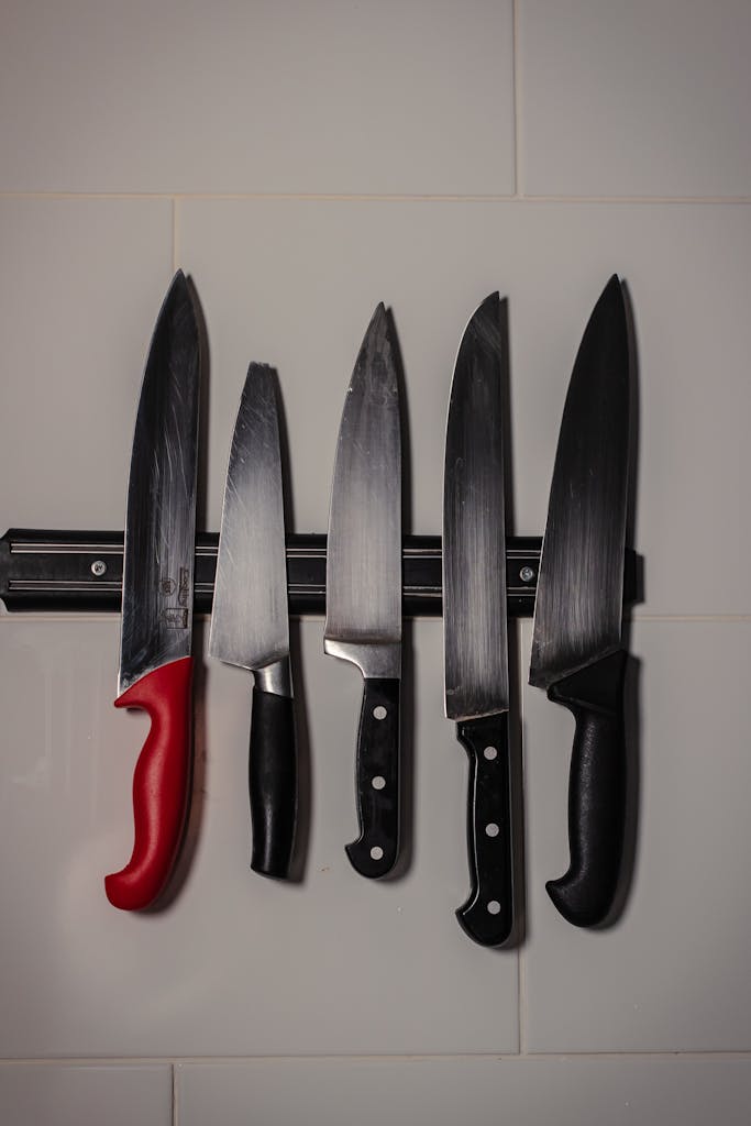 Five kitchen knives hanging on a magnetic strip on a white tiled wall.