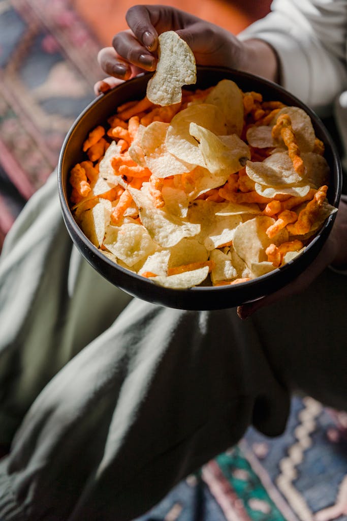 From above of crop anonymous female eating delicious crisps placed in bowl at home