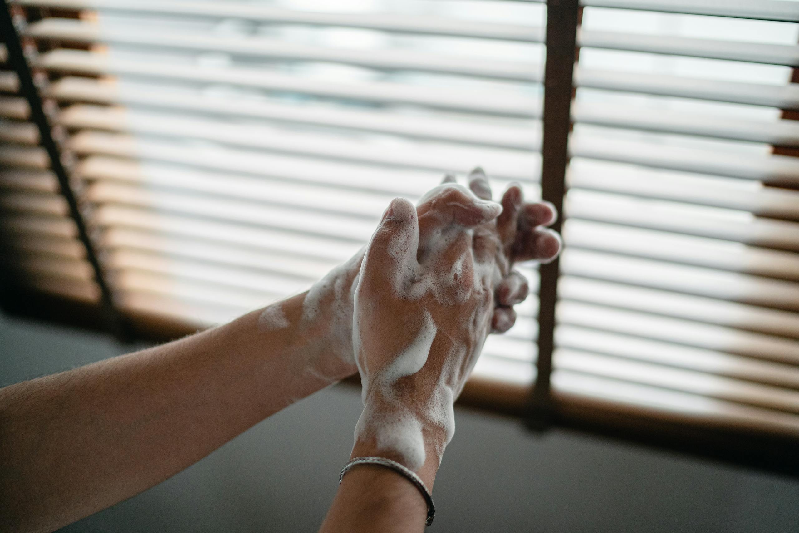 Hands covered in soap lather being washed indoors near a window for hygiene.