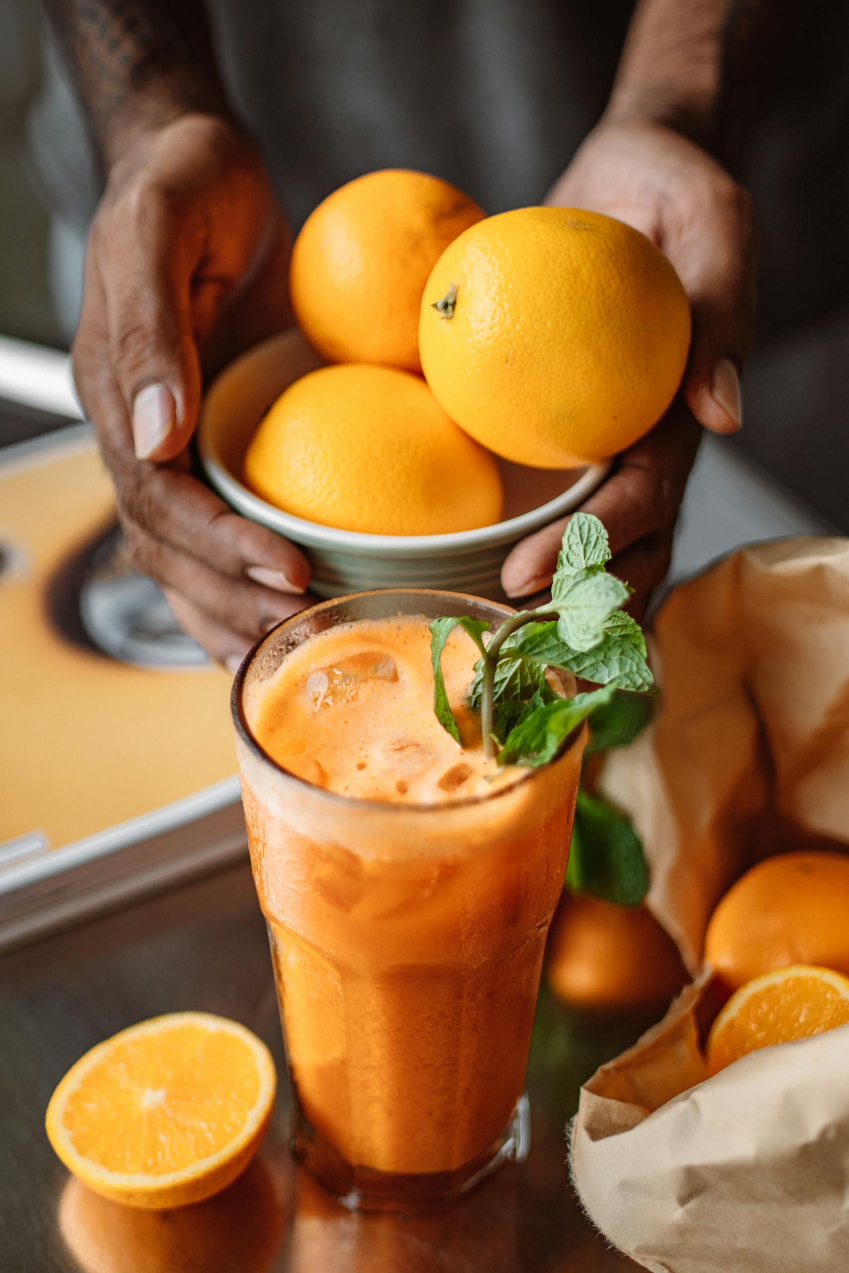 Hands holding bowl of oranges above a freshly made orange juice with mint garnish.