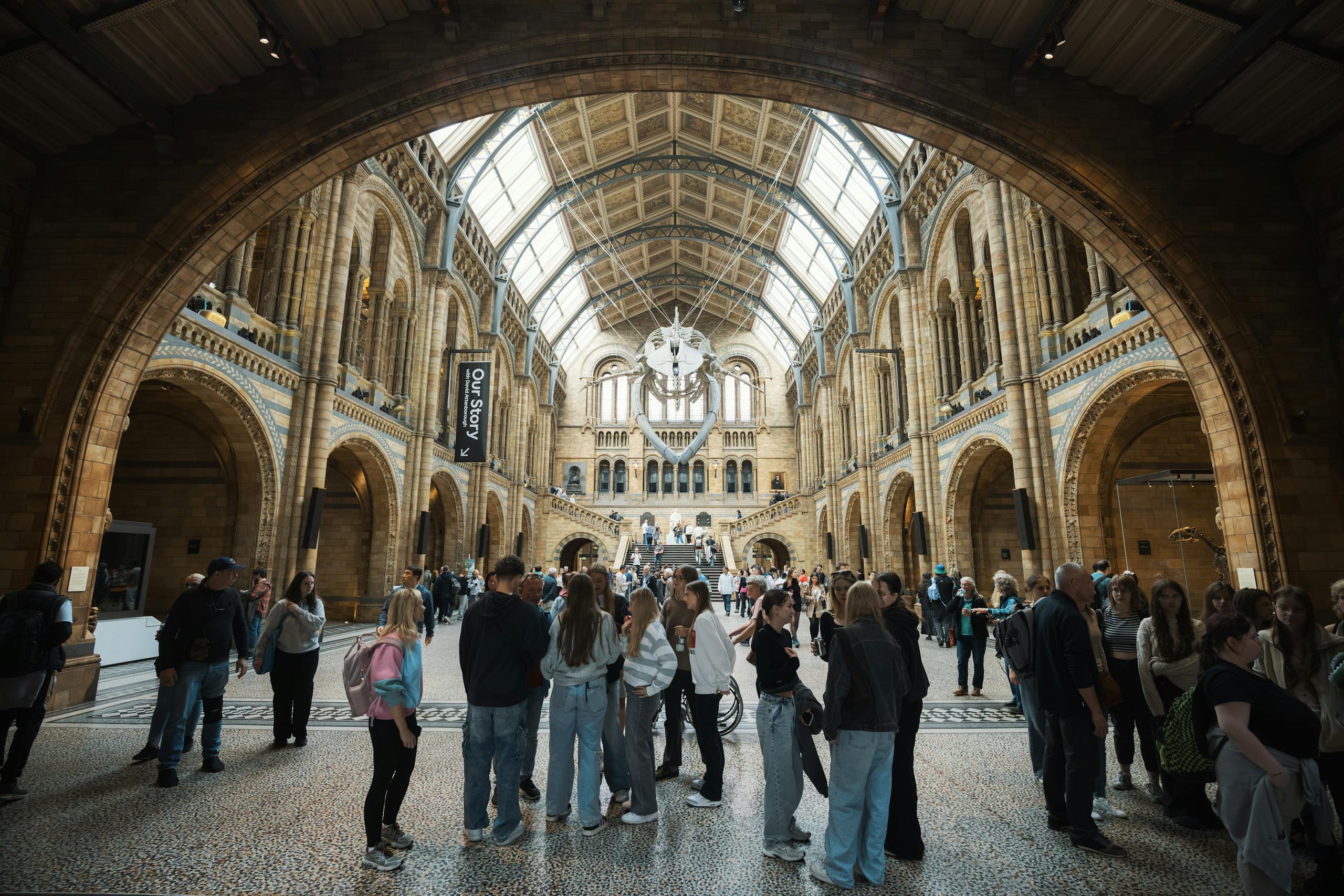 Inside the bustling Natural History Museum with a diverse crowd. Ornate architecture in a bright, dynamic setting.