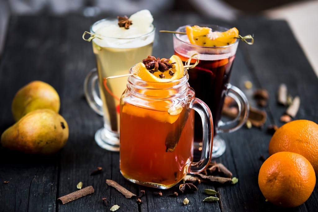 Jar and glasses of mulled wine drinks with fresh orange and pear slices on skewers on table with dry condiments