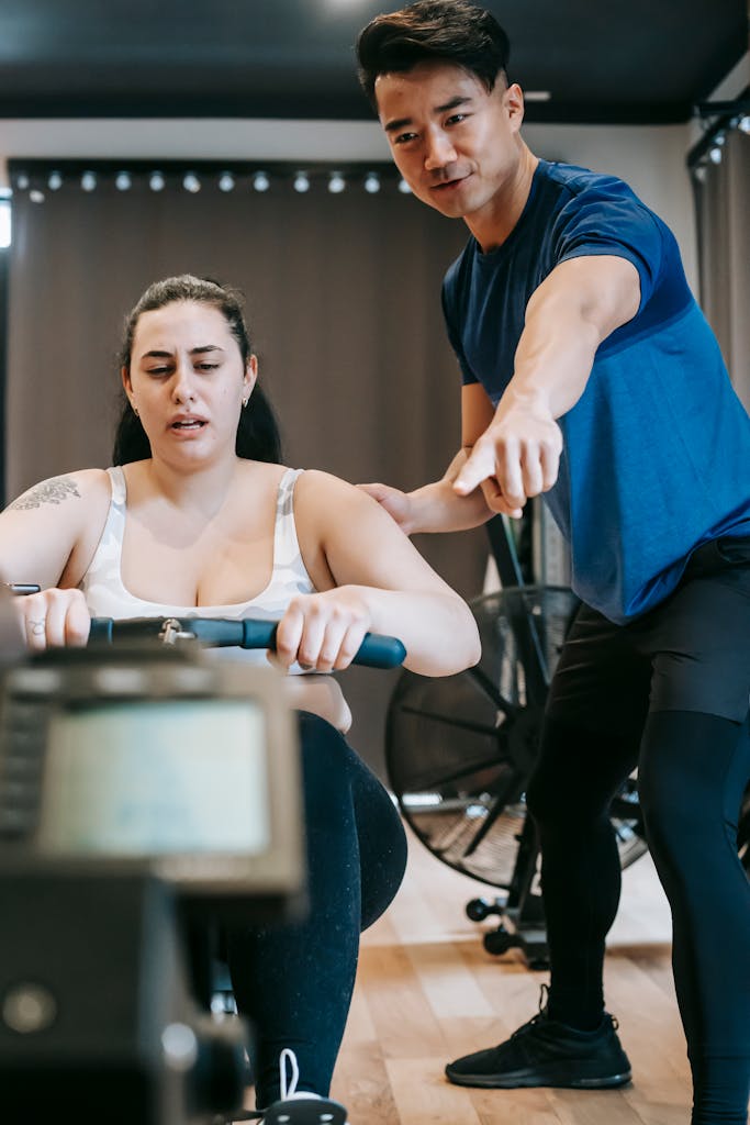 Low angle of Asian male instructor pointing at screen while talking with overweight female on rowing machine