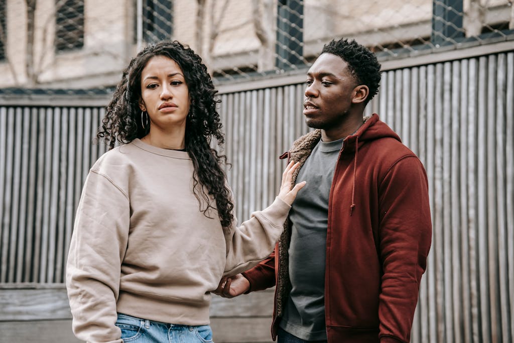 Sad ethnic girlfriend with curly hair rejecting annoyed African American boyfriend while arguing on street near wooden fence during breakup