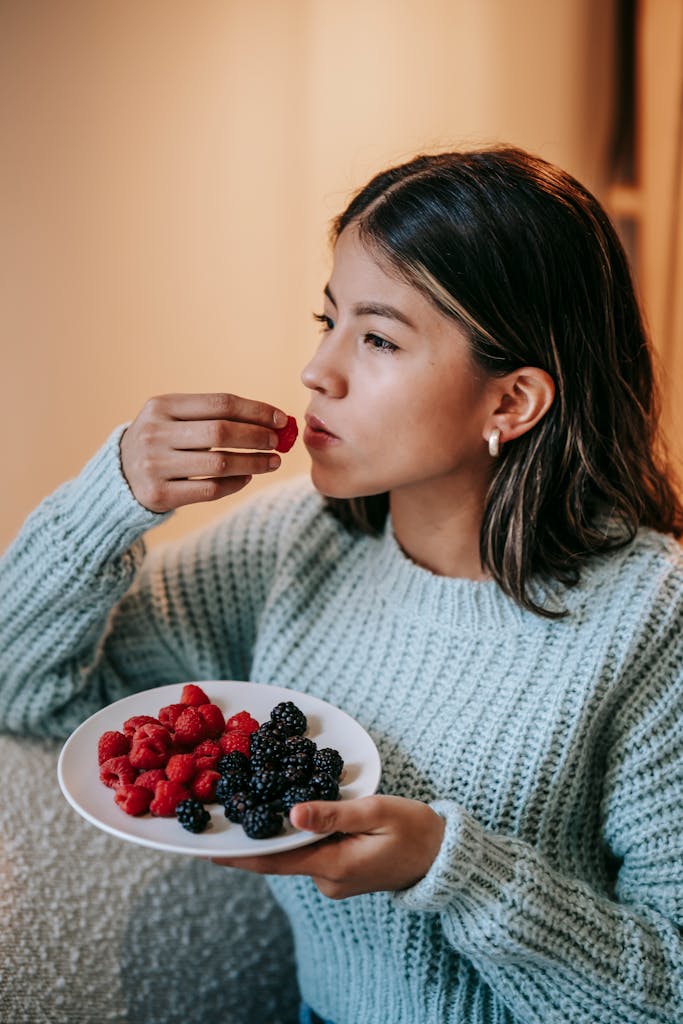 Serious Asian woman in sweater sitting on sofa with plate and eating yummy raspberry at home