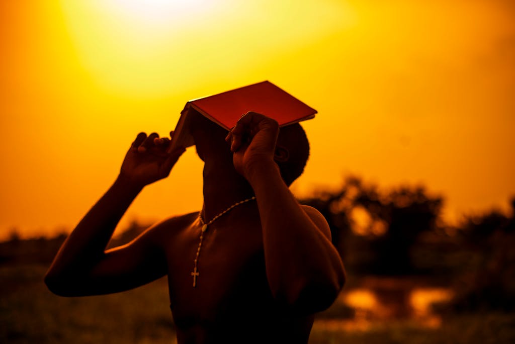 Shirtless man holding a book, silhouetted against a vibrant sunset. Powerful symbolism and serene atmosphere.
