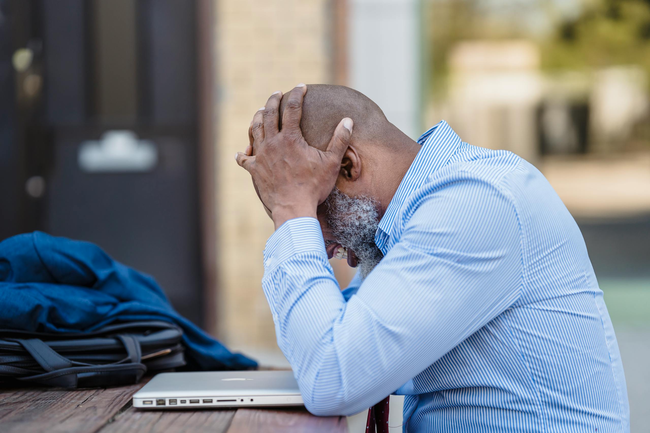 Side view of pensive middle aged African American male entrepreneur wearing blue shirt grabbing head with hands while sitting at table with closed laptop in veranda