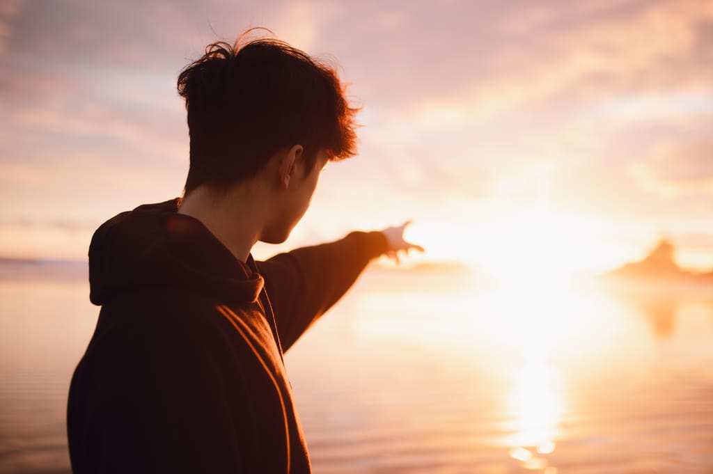 Silhouette of a man pointing towards a sunrise over the sea, casting a warm glow.