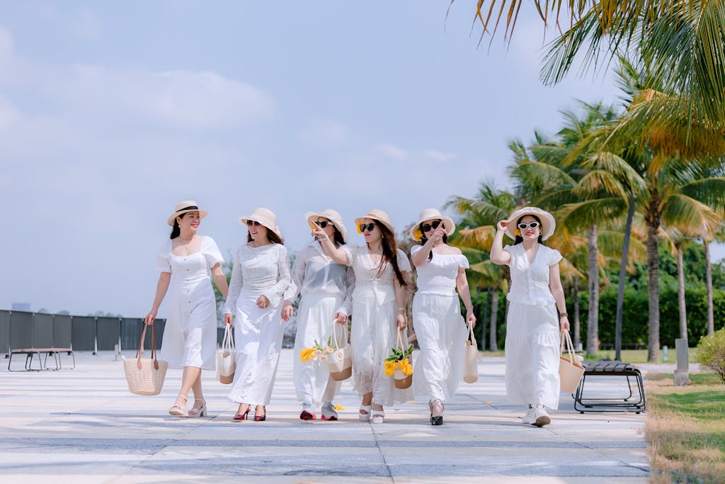 Six women in white dresses and hats walk in the sun, carrying bags, surrounded by palm trees.
