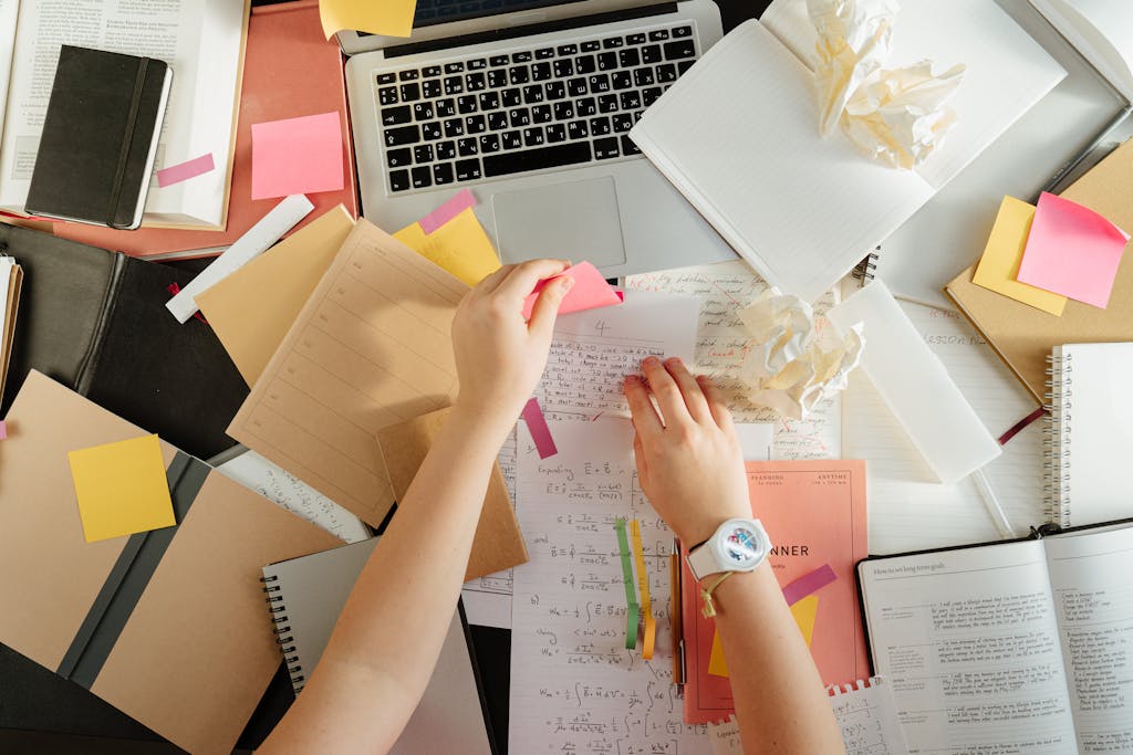 Top view of a workspace with books, a laptop, and hands organizing notes using sticky notes.