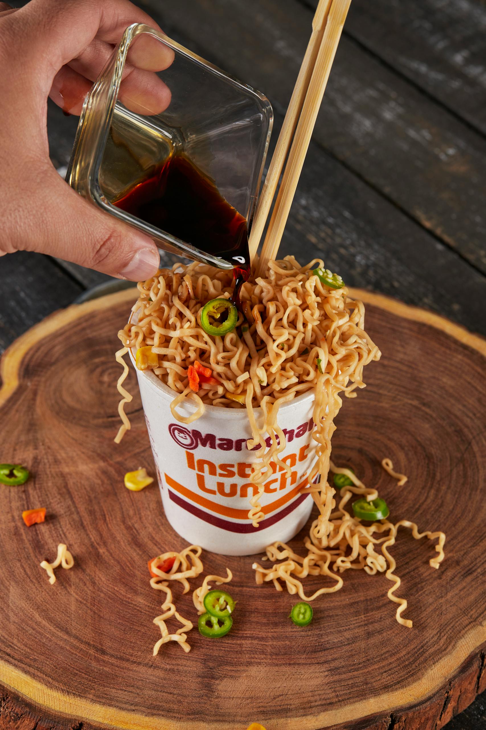 Top view of instant noodles being flavored with soy sauce, featuring chopsticks on a wooden table.