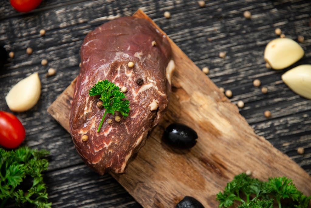Top view of raw beef steak garnished with peppercorns and parsley on a wooden board with garlic and olives.