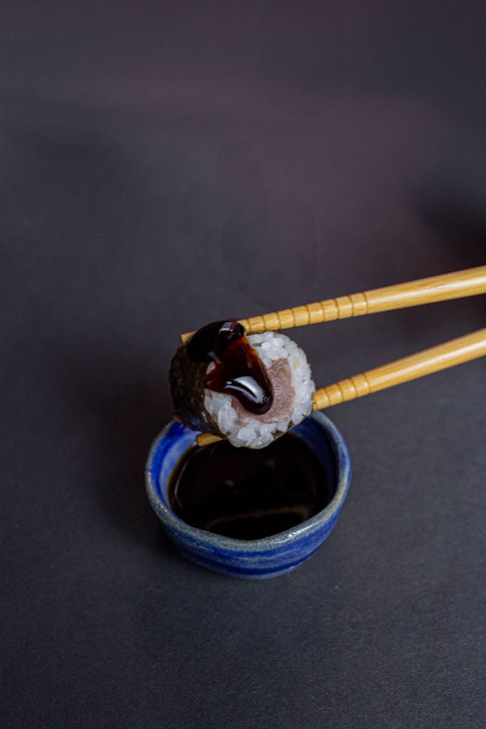 Top view of sushi roll held with chopsticks over a soy sauce bowl on a dark background.