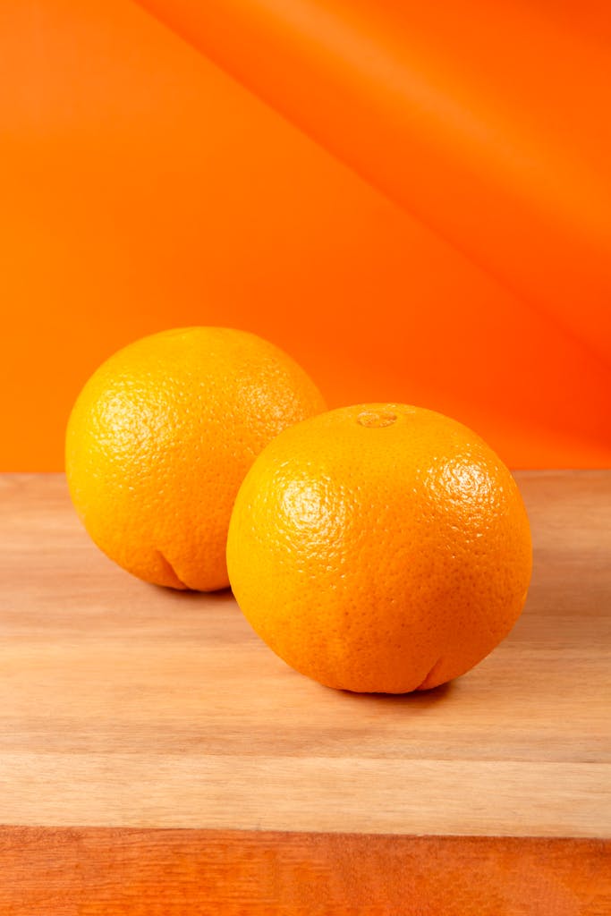 Two fresh oranges on a wooden surface with vibrant orange background, showcasing their bright, juicy texture.