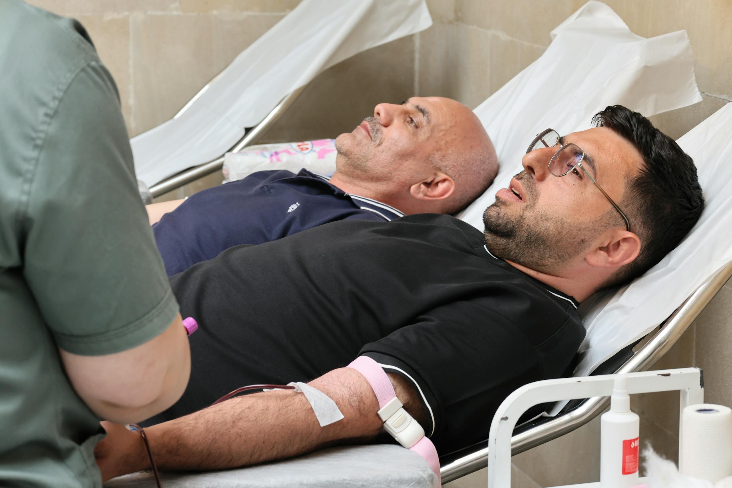 Two men are lying on medical beds while donating blood at a clinic.