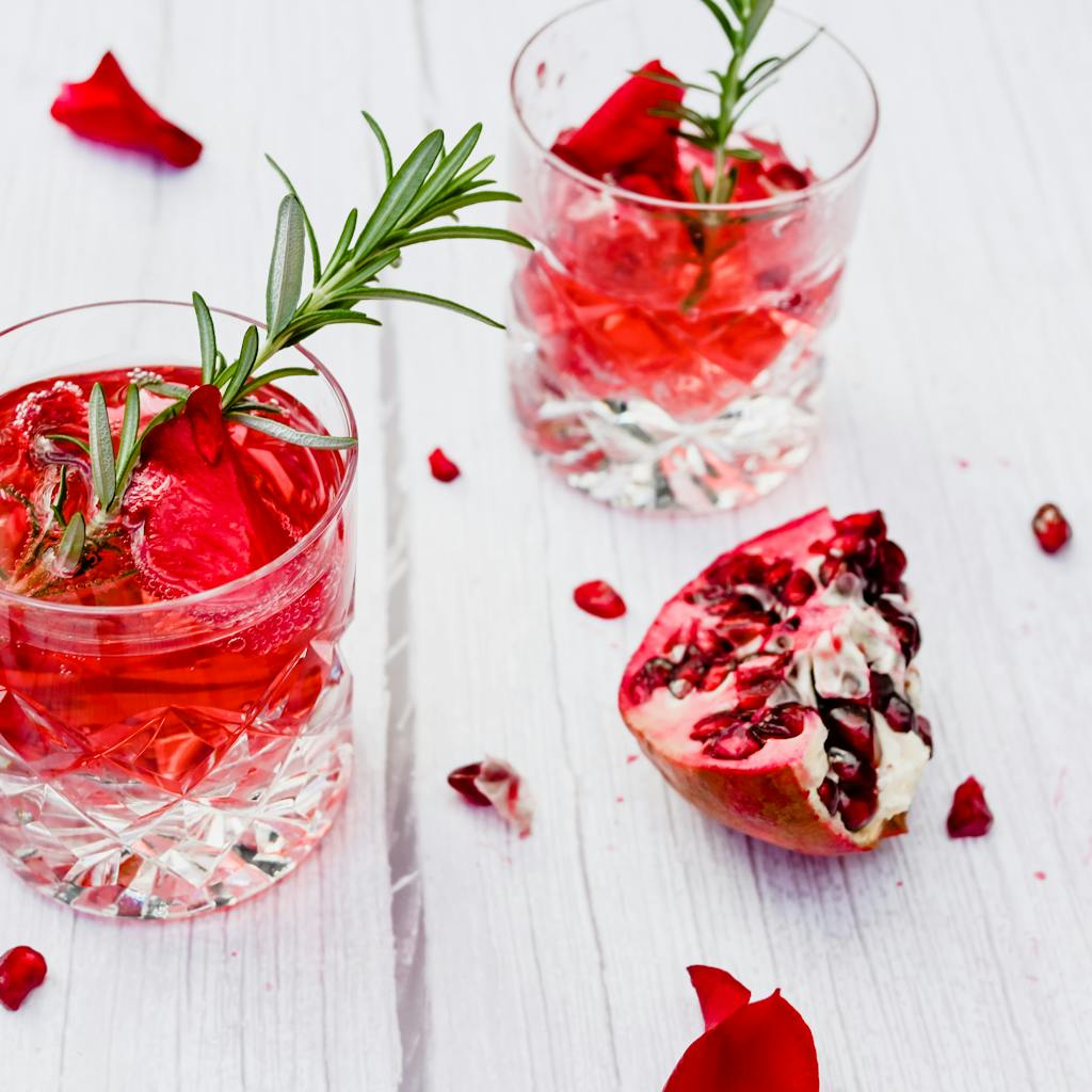 Two pomegranate rosemary cocktails in crystal glasses with fresh garnish on a white wooden table.