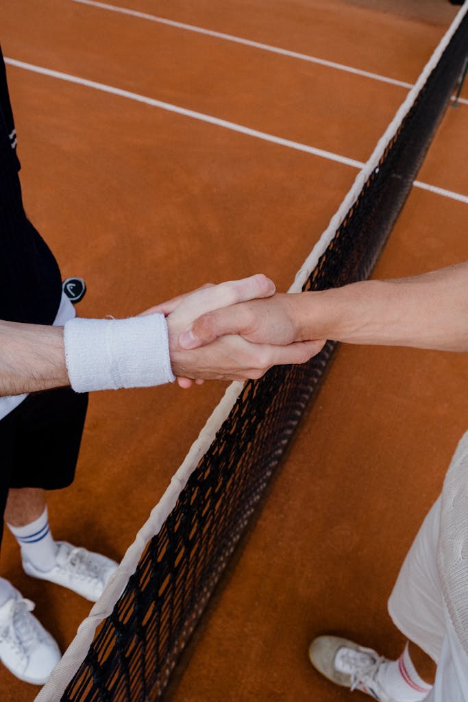 Two tennis players in a friendly handshake over a clay tennis court net, symbolizing sportsmanship.